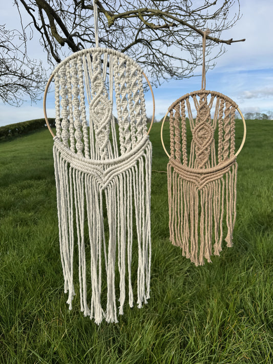Two macrame wall hangings hanging outdoors on a grassy area with trees in the background.