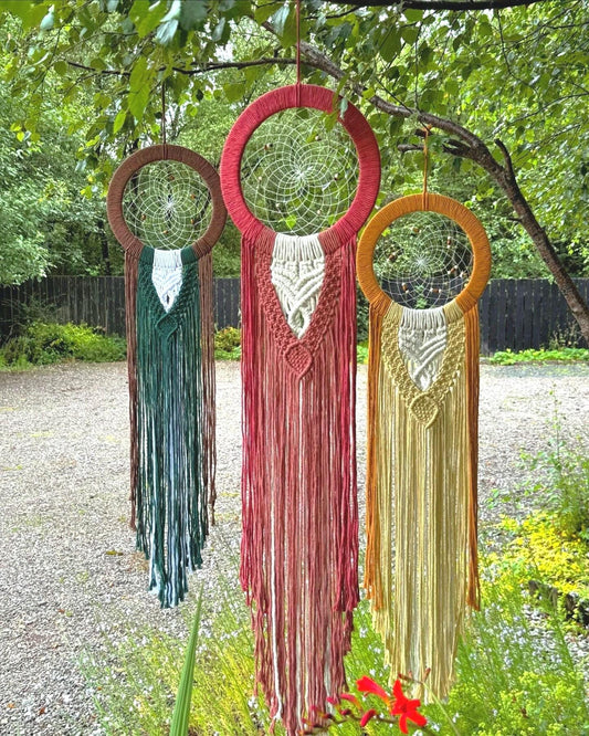 Three colourful macrame dream catchers hanging outdoors with greenery in the background.