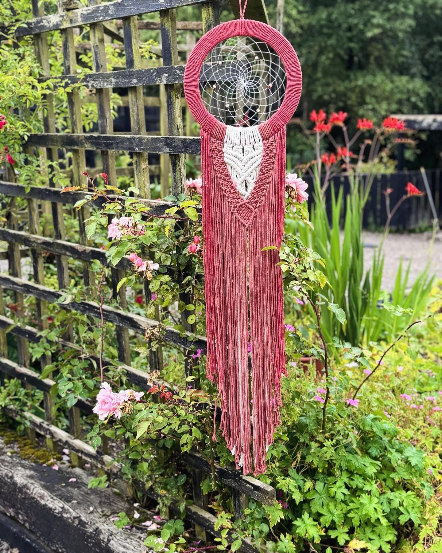 Red macrame wall hanging with a dreamcatcher design in a garden setting