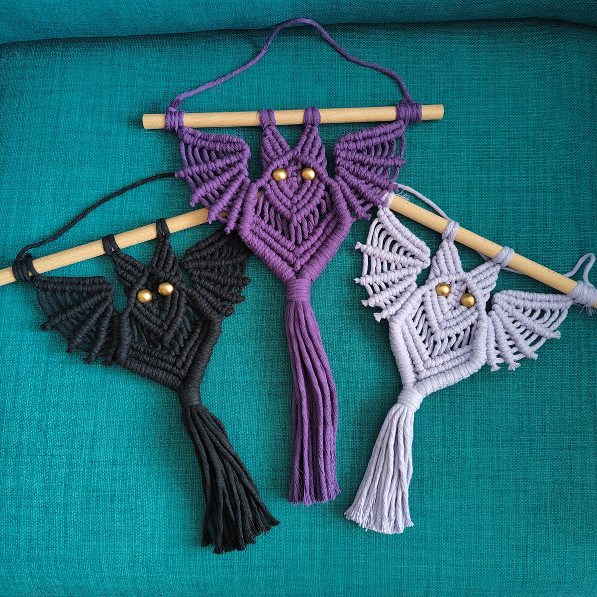 Three macrame bat wall hangings in black, purple, and light lavender, each attached to a wooden dowel and displayed on a teal textured fabric background.