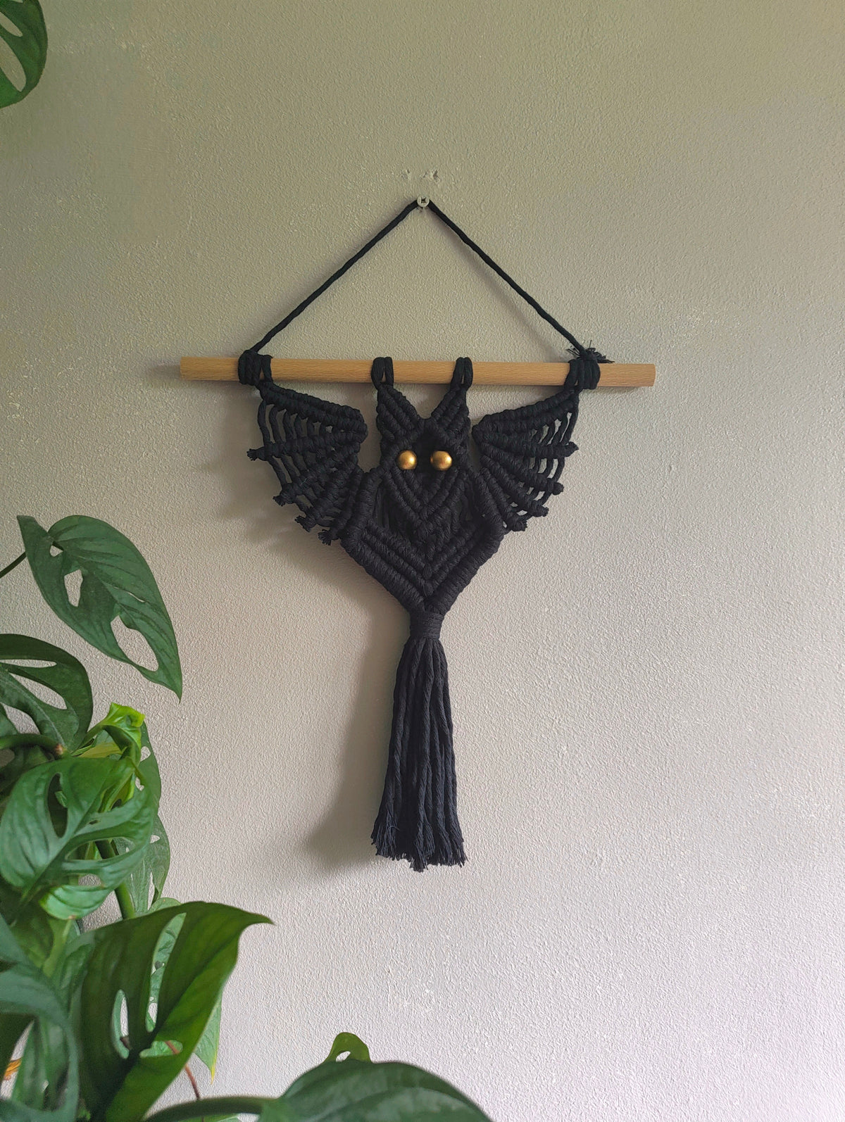 A black macrame bat wall hanging suspended from a light wooden dowel, with small gold bead eyes, displayed on a textured beige wall with green plant leaves in the foreground.