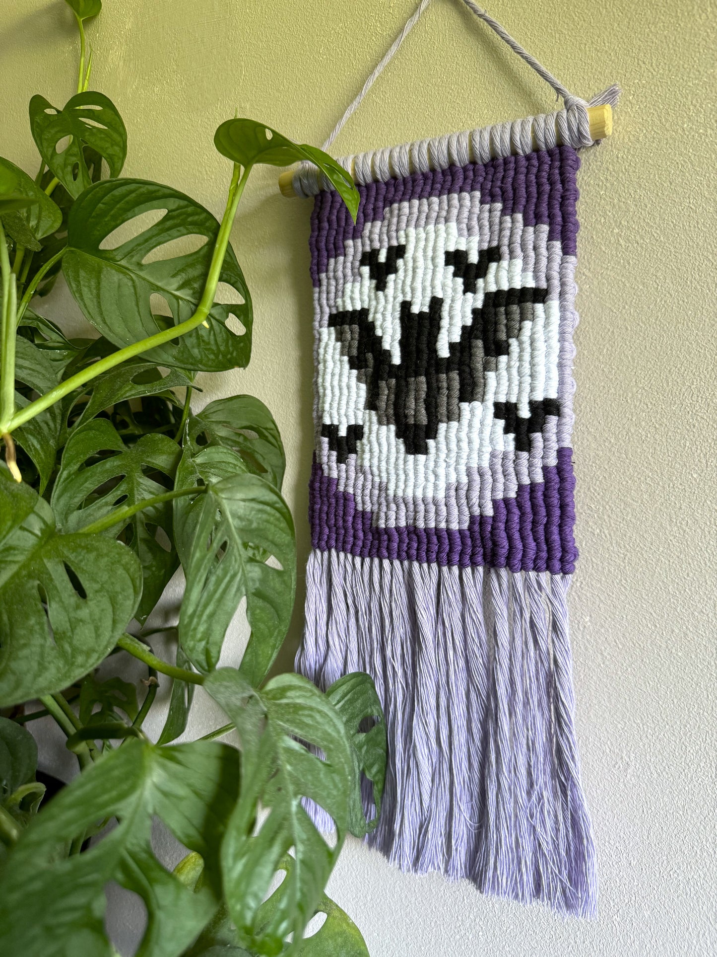 Handmade purple macrame wall hanging with a pixelated bats design on a light green wall next to a plant.