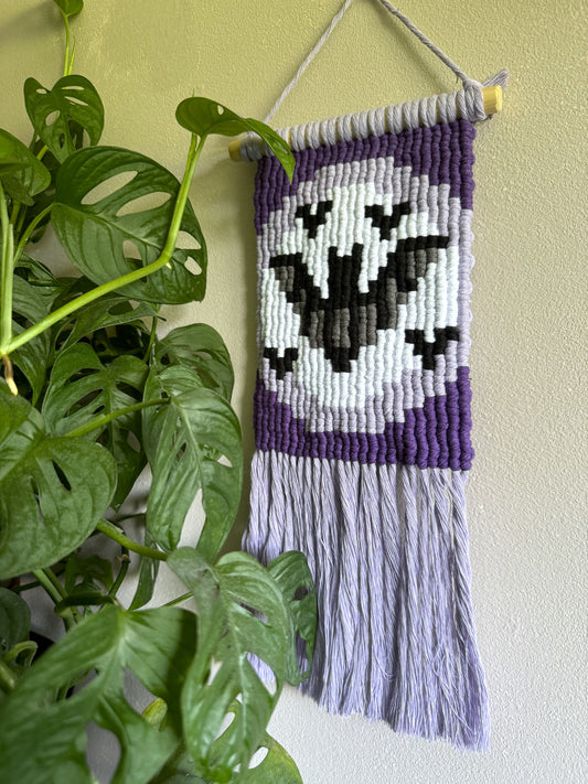 Handmade purple macrame wall hanging with a pixelated bats design on a light green wall next to a plant.