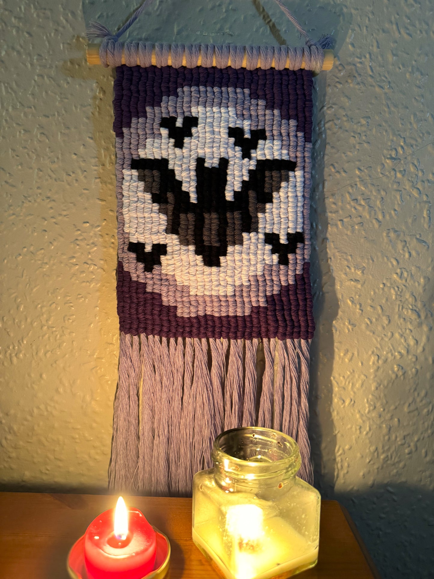 Pixel Macrame wall hanging with bats design on a textured wall, lit by candles.