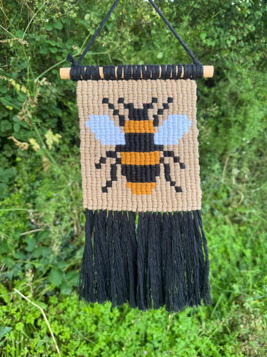 Handmade wall hanging with a bee design and black fringes, hanging against a natural green background.