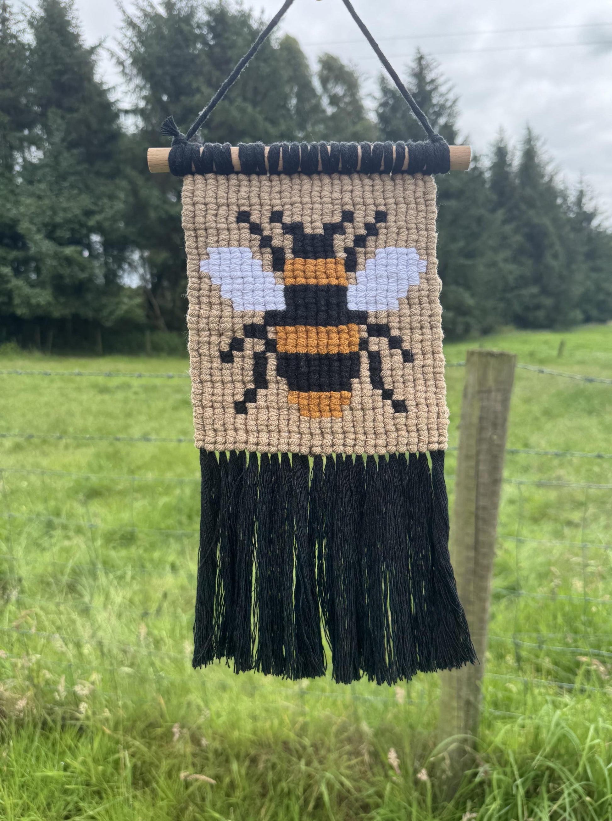 Woven wall hanging with a bee design and black fringes, hanging outdoors.