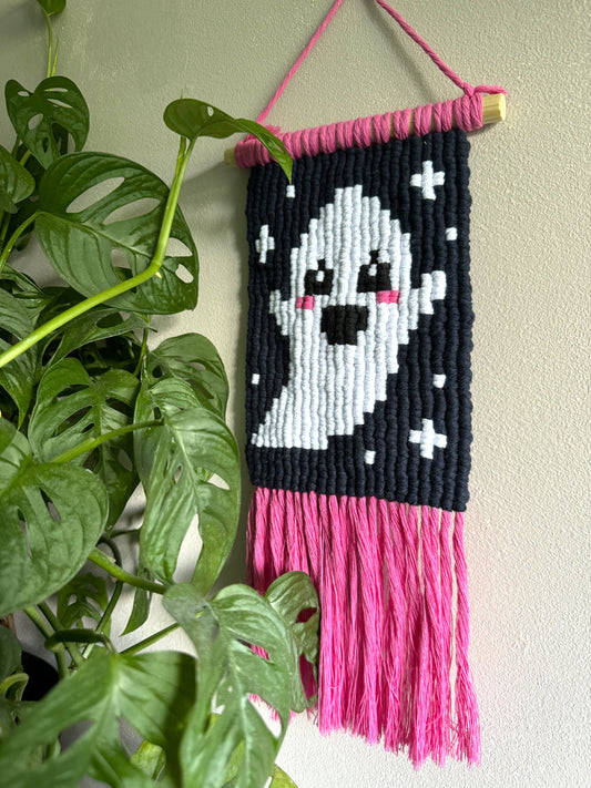 Handmade macrame wall hanging with pixelated ghost design and pink fringe, hanging on a wall next to a plant.