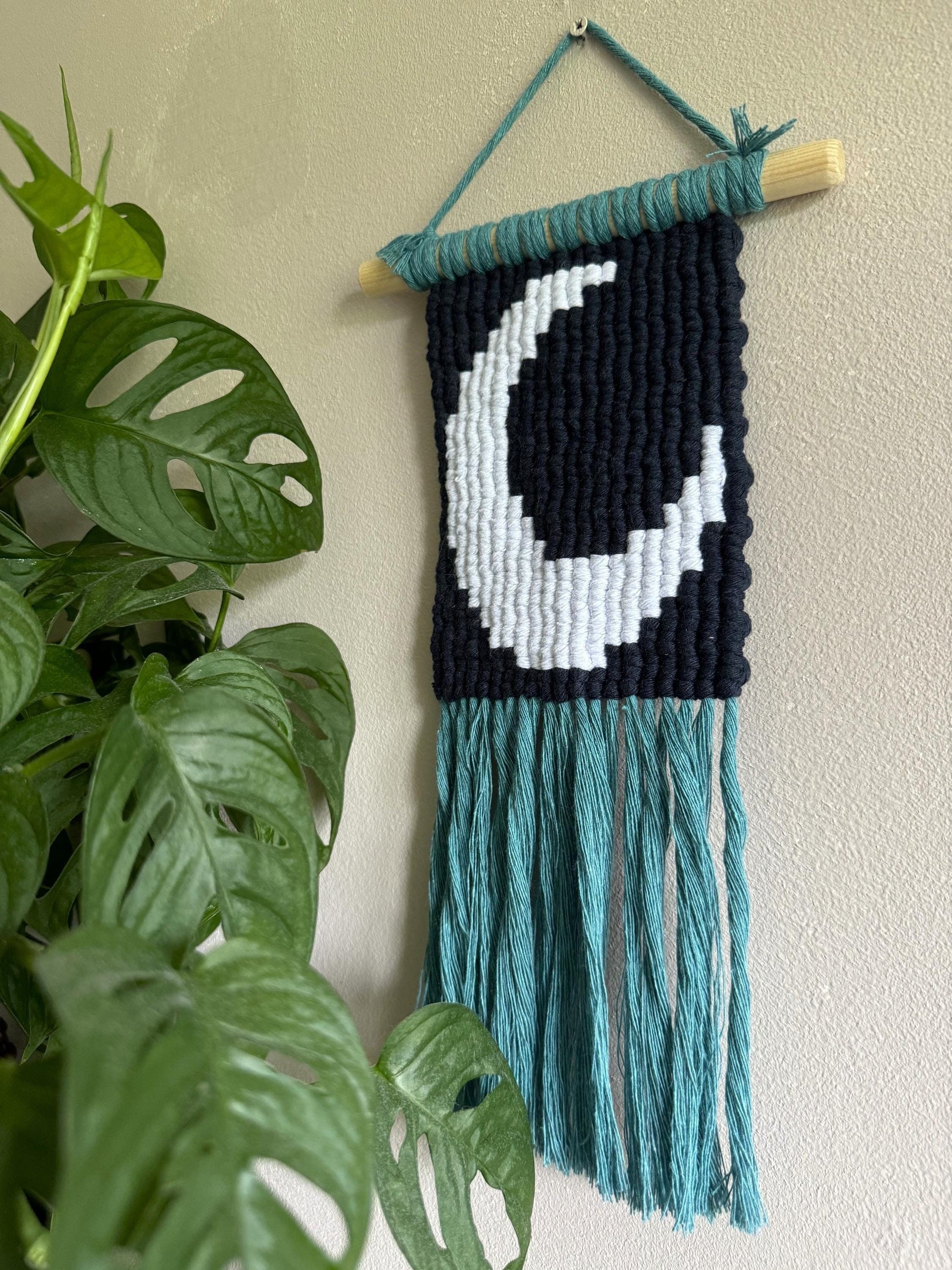Macrame wall hanging with a moon design and teal fringe next to a plant on a beige wall.