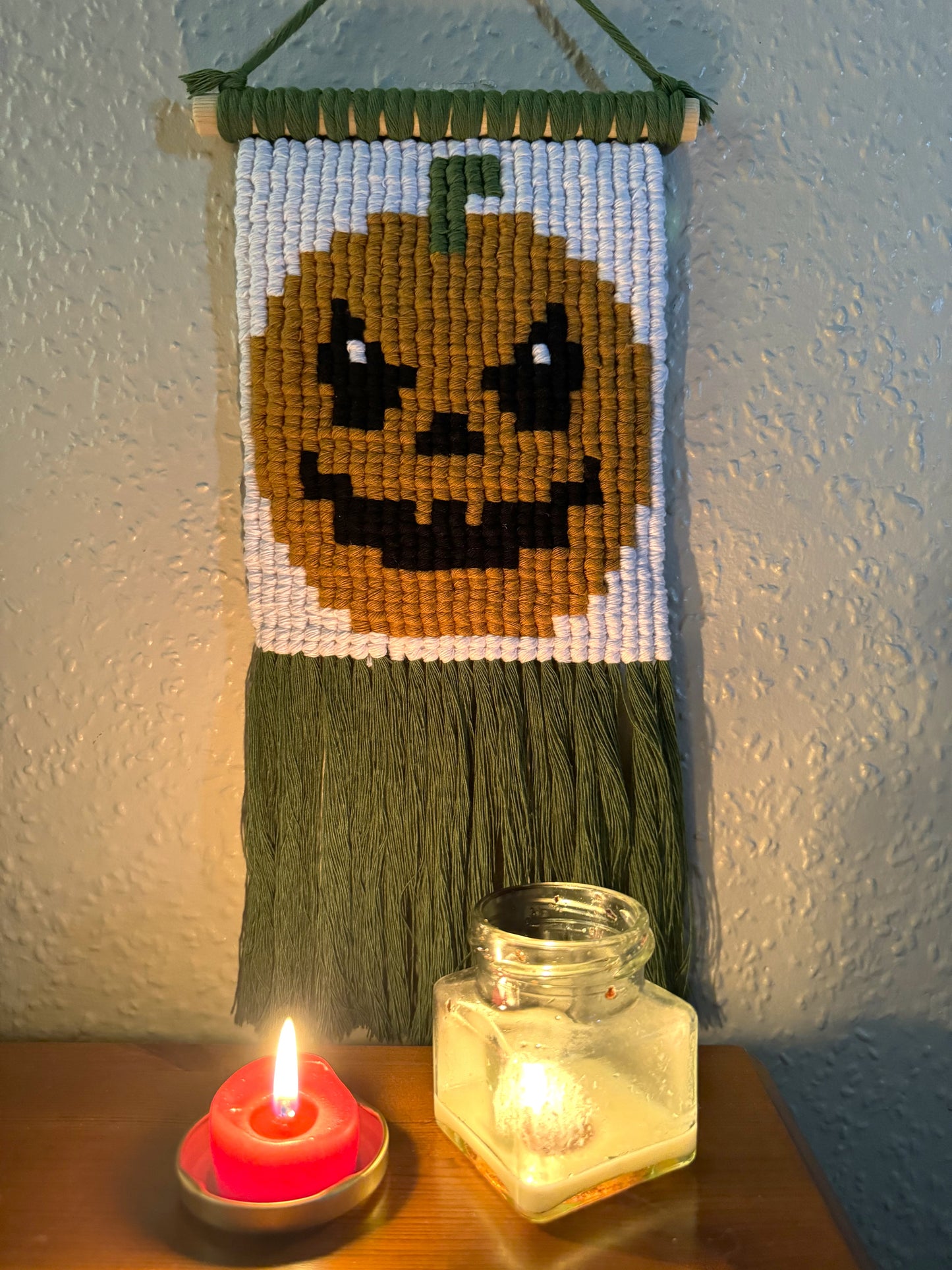 Pixelated pumpkin design on a macrame wall hanging with candles below.