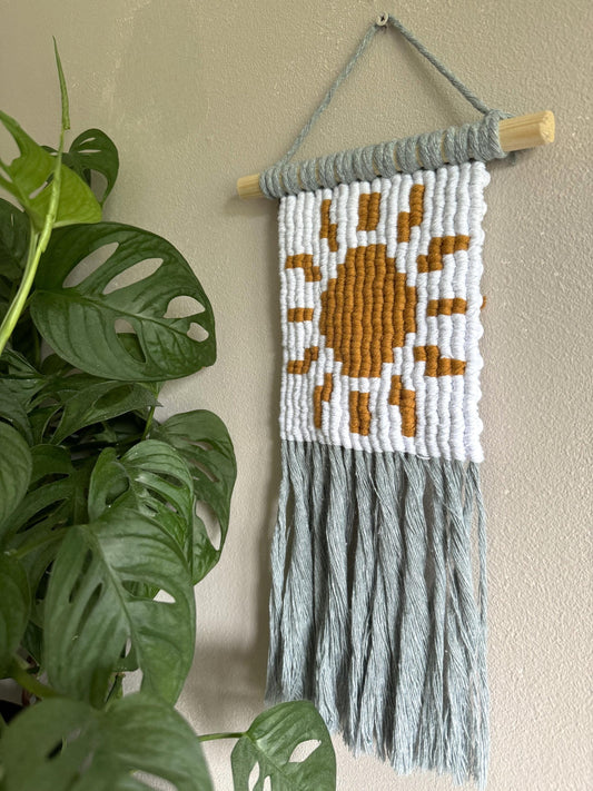 Macrame wall hanging with sun design and fringe, next to a plant on a beige wall.