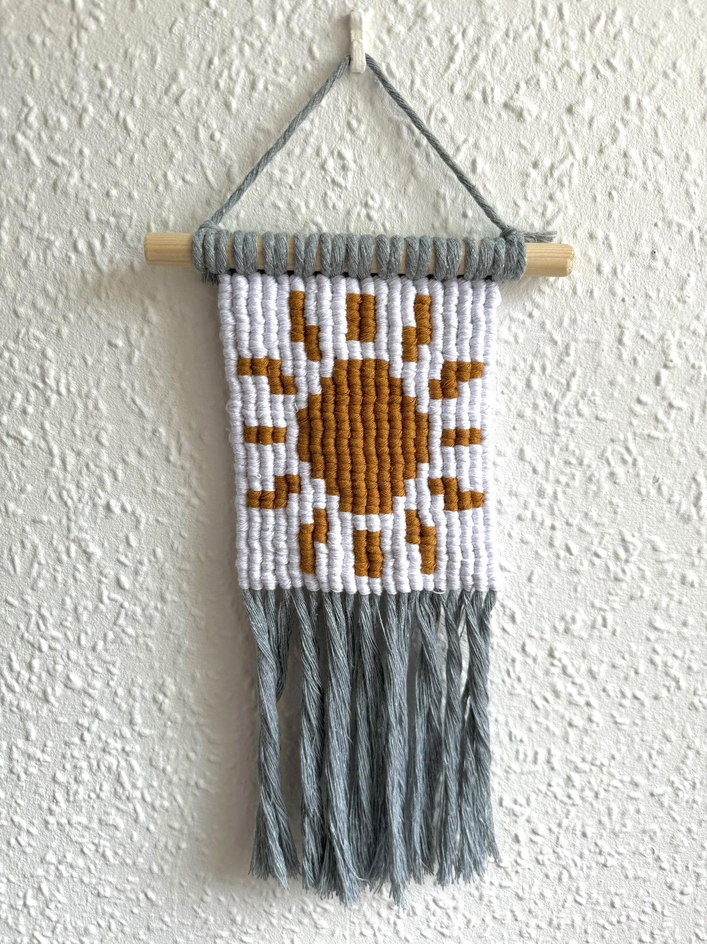 Pixel macrame wall hanging with sun design and gray fringe on a white wall