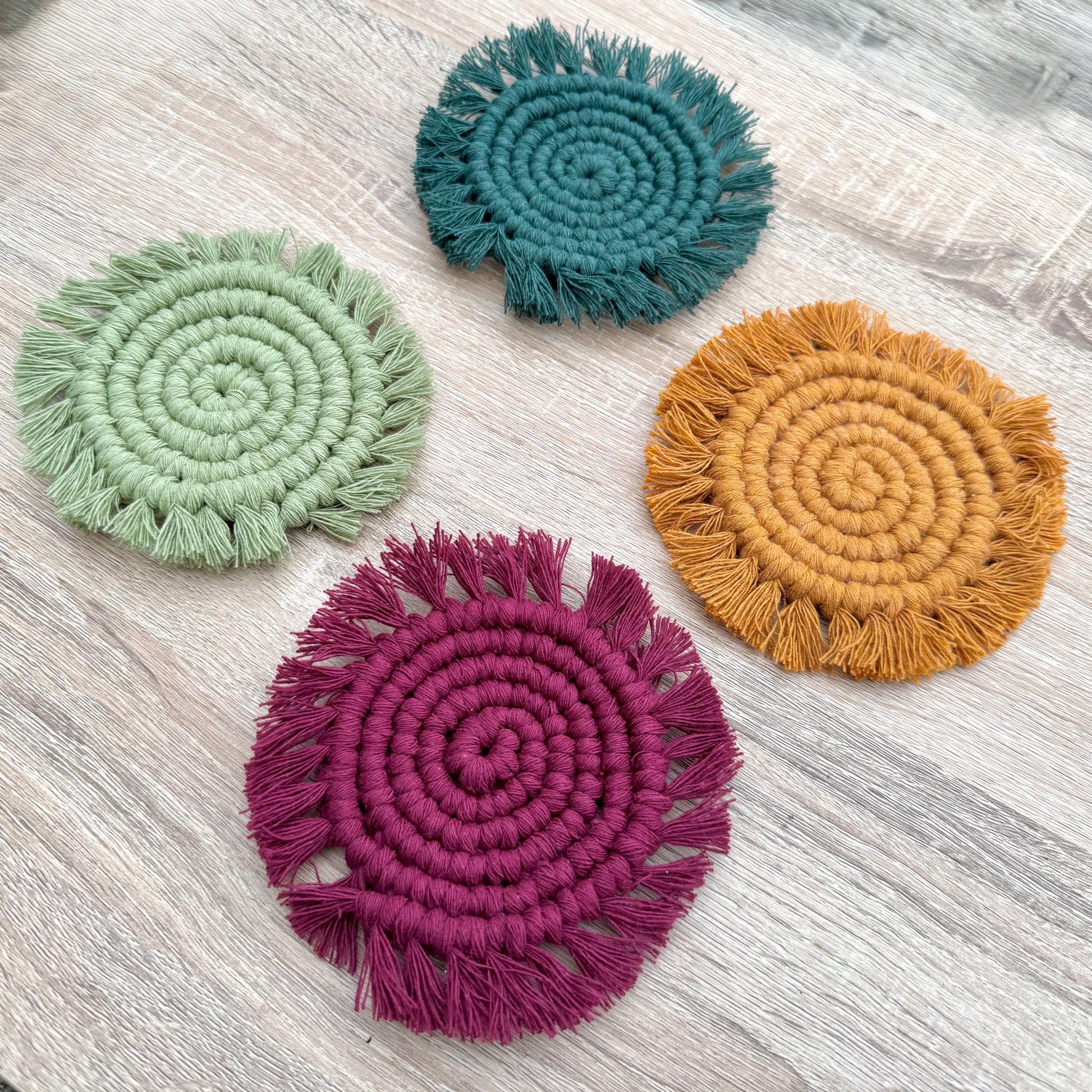 Four round woven coasters in light green, dark green, yellow and burgundy on a light wooden surface.
