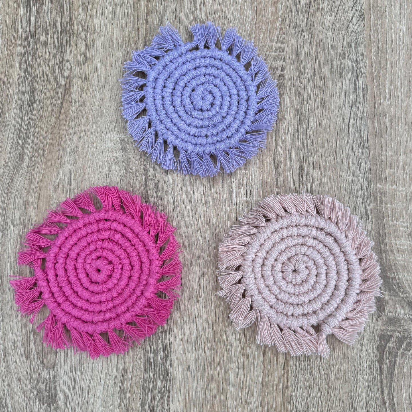 Three circular macrame coasters in colours bright pink, lilac and light pink with tassels on a wooden surface.