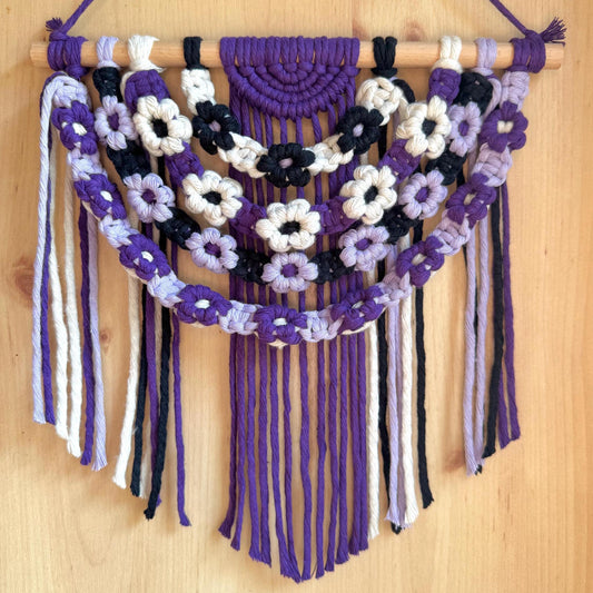 Macrame wall hanging with floral patterns in purple, white, and black on a wooden background
