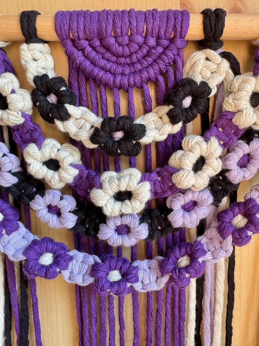 Macrame wall hanging with floral patterns in purple, black, white, and beige on a wooden rod.