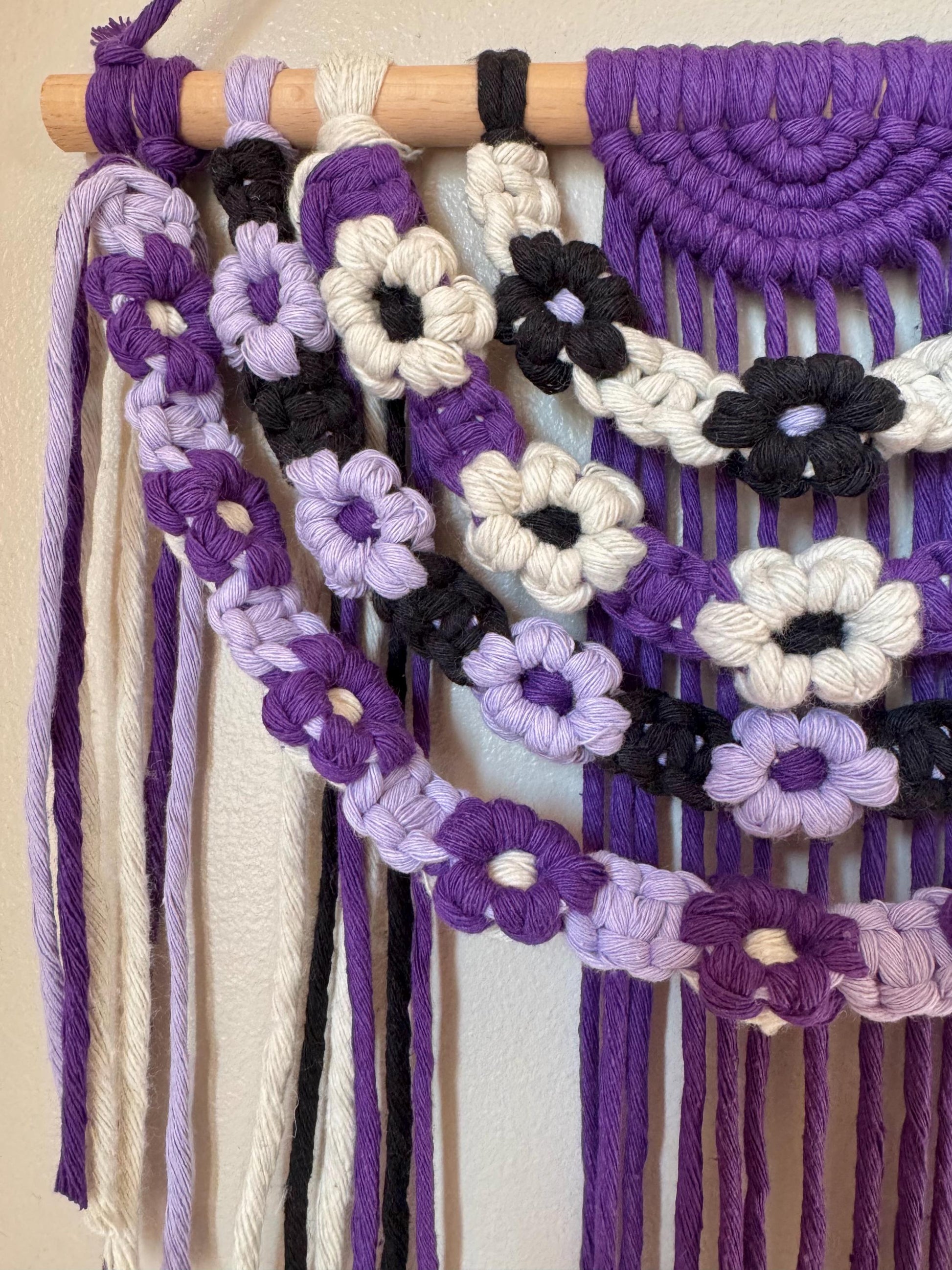 Macrame wall hanging with purple, white, and black flower patterns on a beige background