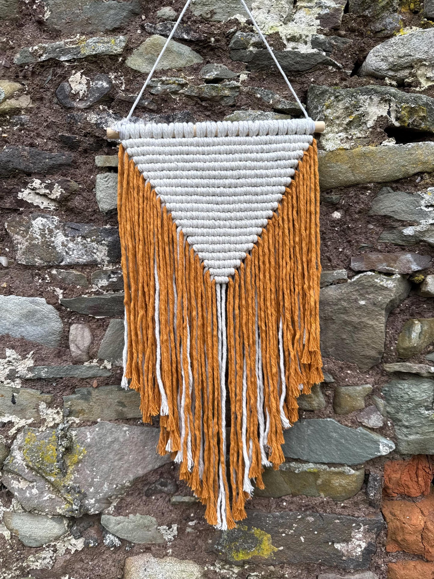 Woven wall hanging with mustard yellow and white fringes against a stone wall.