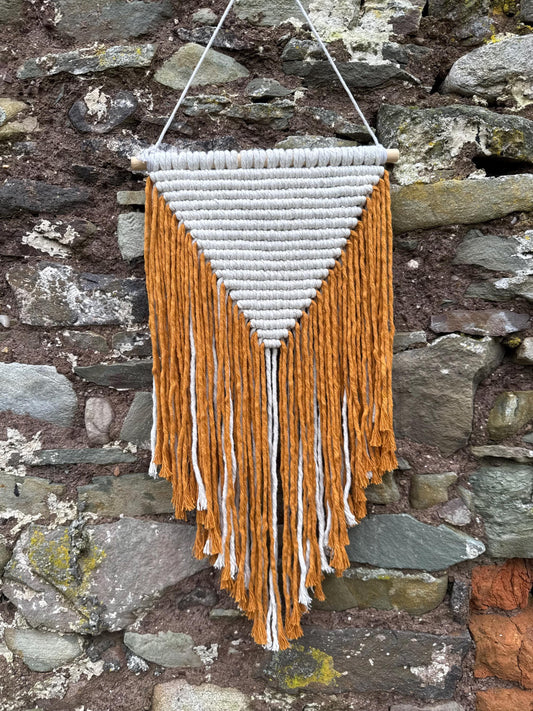 Woven wall hanging with mustard yellow and white fringes against a stone wall.