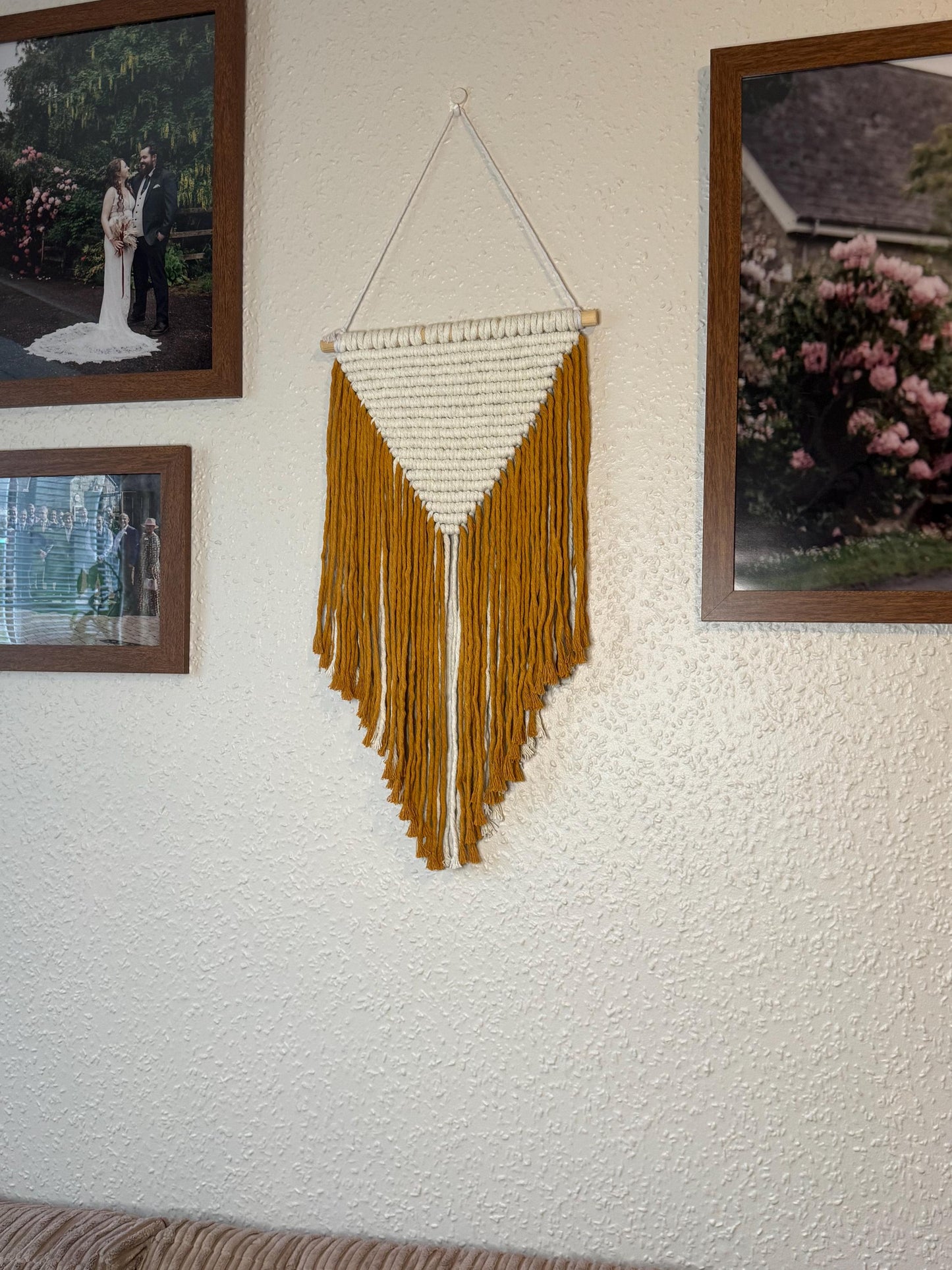 Yellow macrame wall hanging with fringes on a textured wall with framed photographs.
